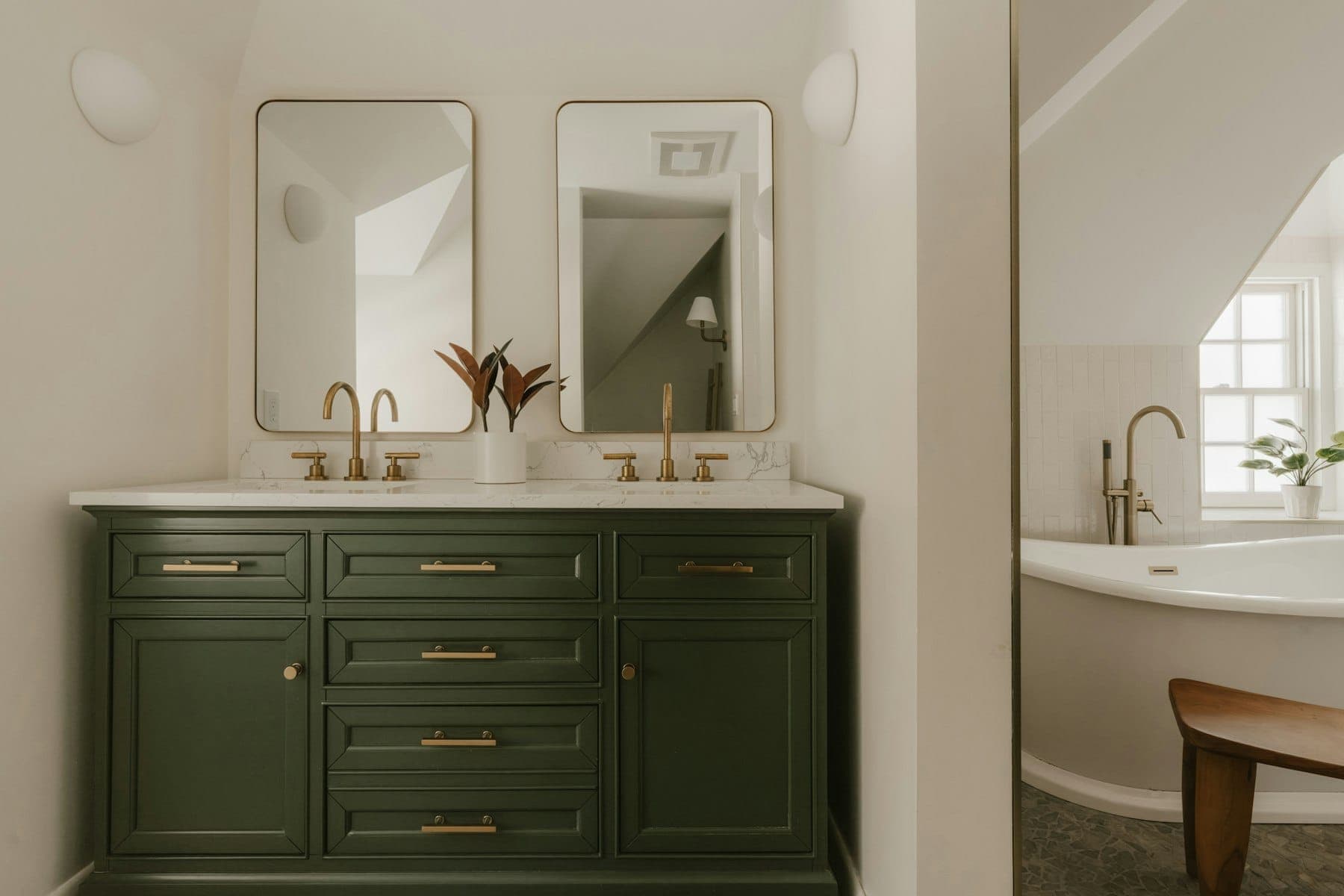 Green double vanity with brass faucets and rounded mirrors beside a freestanding tub under a sloped ceiling
