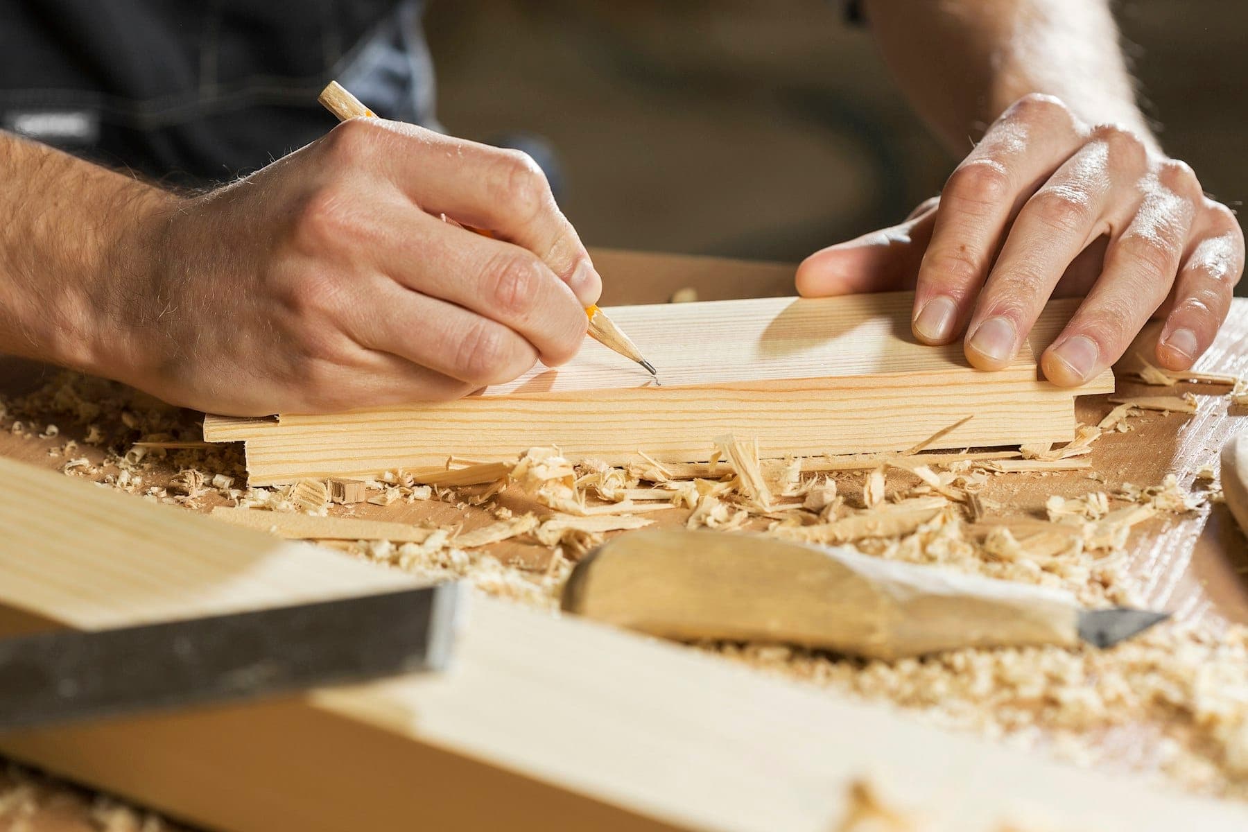Hands marking a cut line with a carpenter's pencil on a pine board, with a chisel and wood shavings nearby