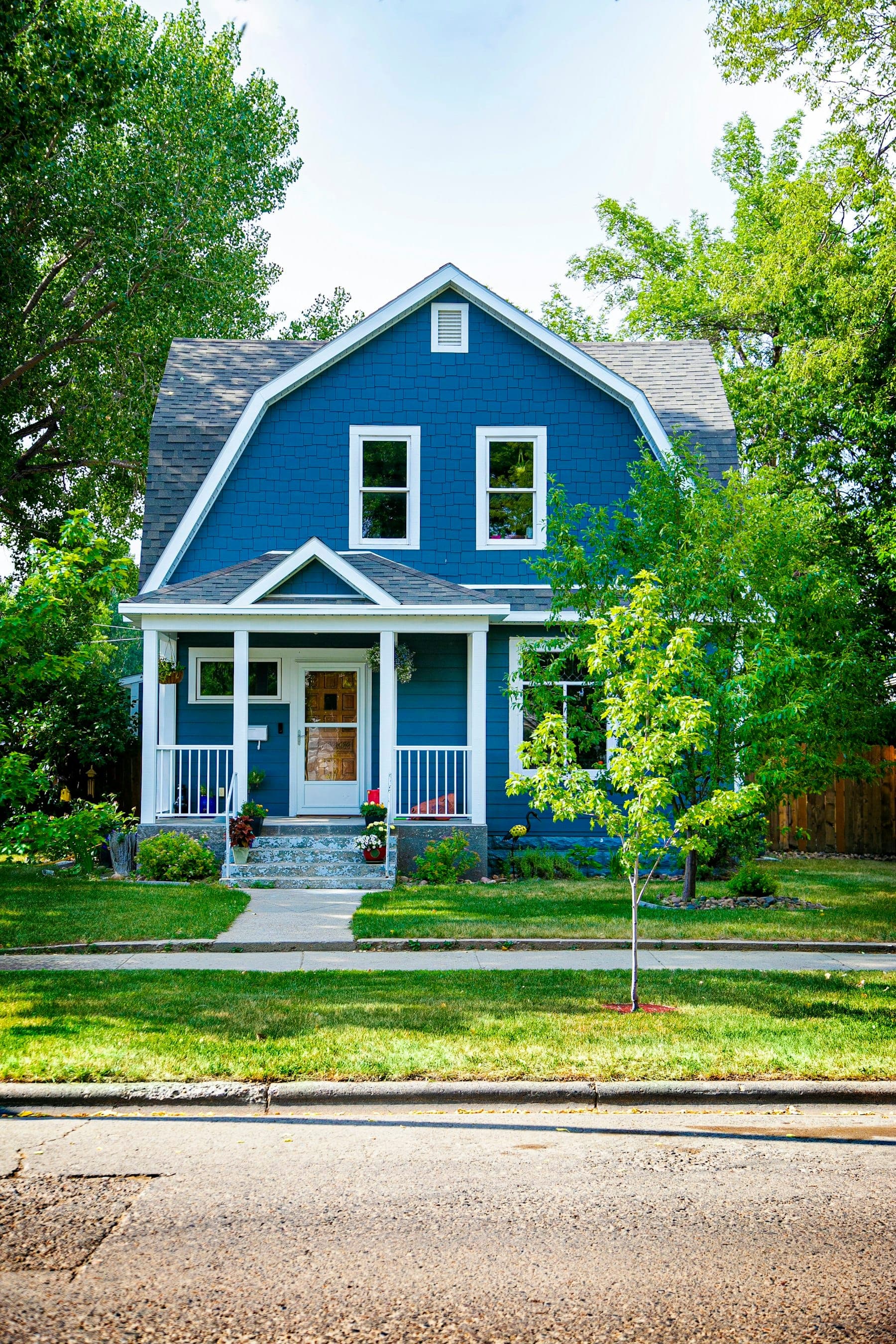 Blue Dutch colonial house with crisp white trim, a covered front porch and a young tree in the lawn
