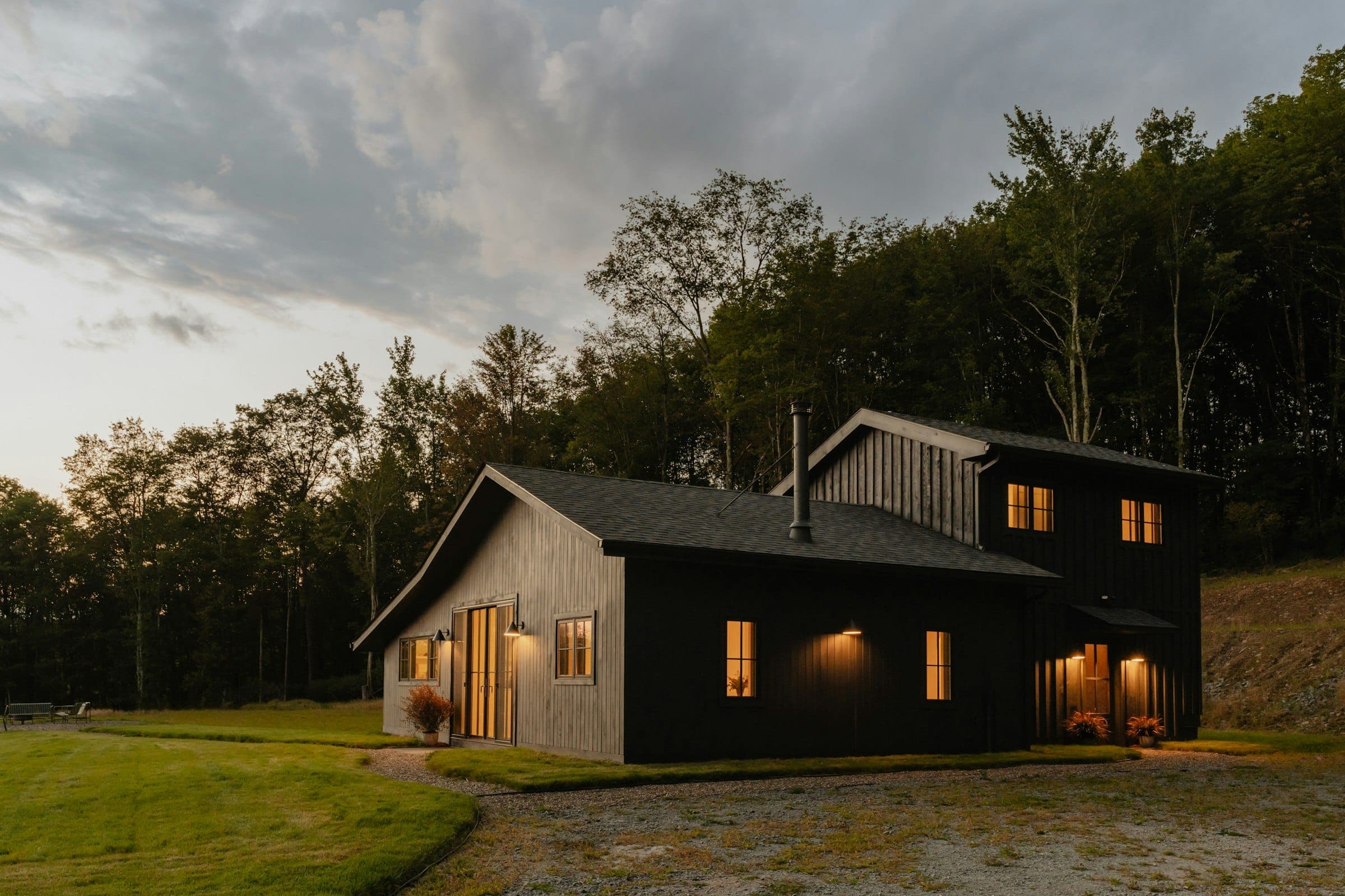 Black board-and-batten farmhouse with warm-lit windows at dusk, set against a wooded hillside