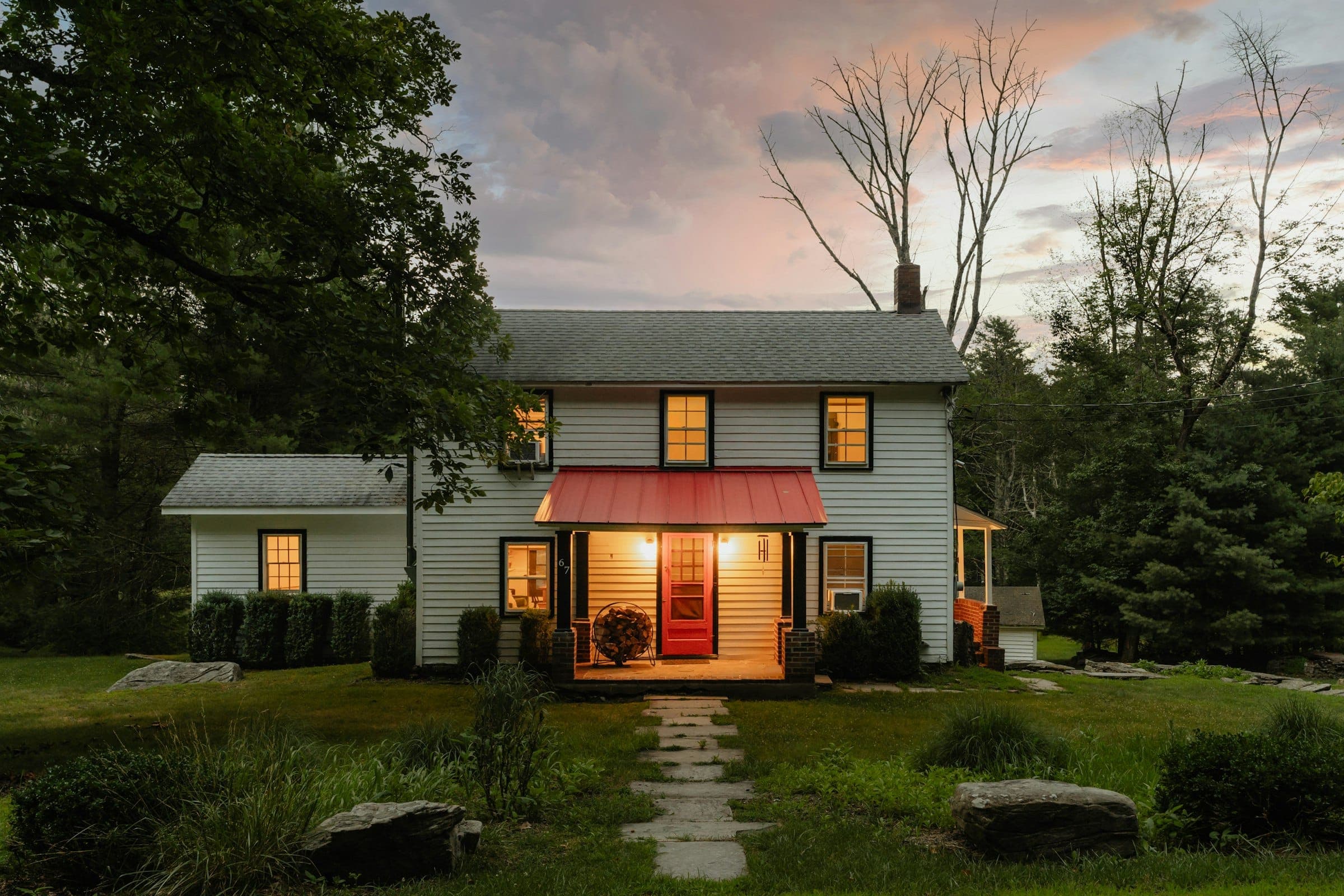 White clapboard farmhouse at dusk with lit windows and a glowing front porch under a red metal awning