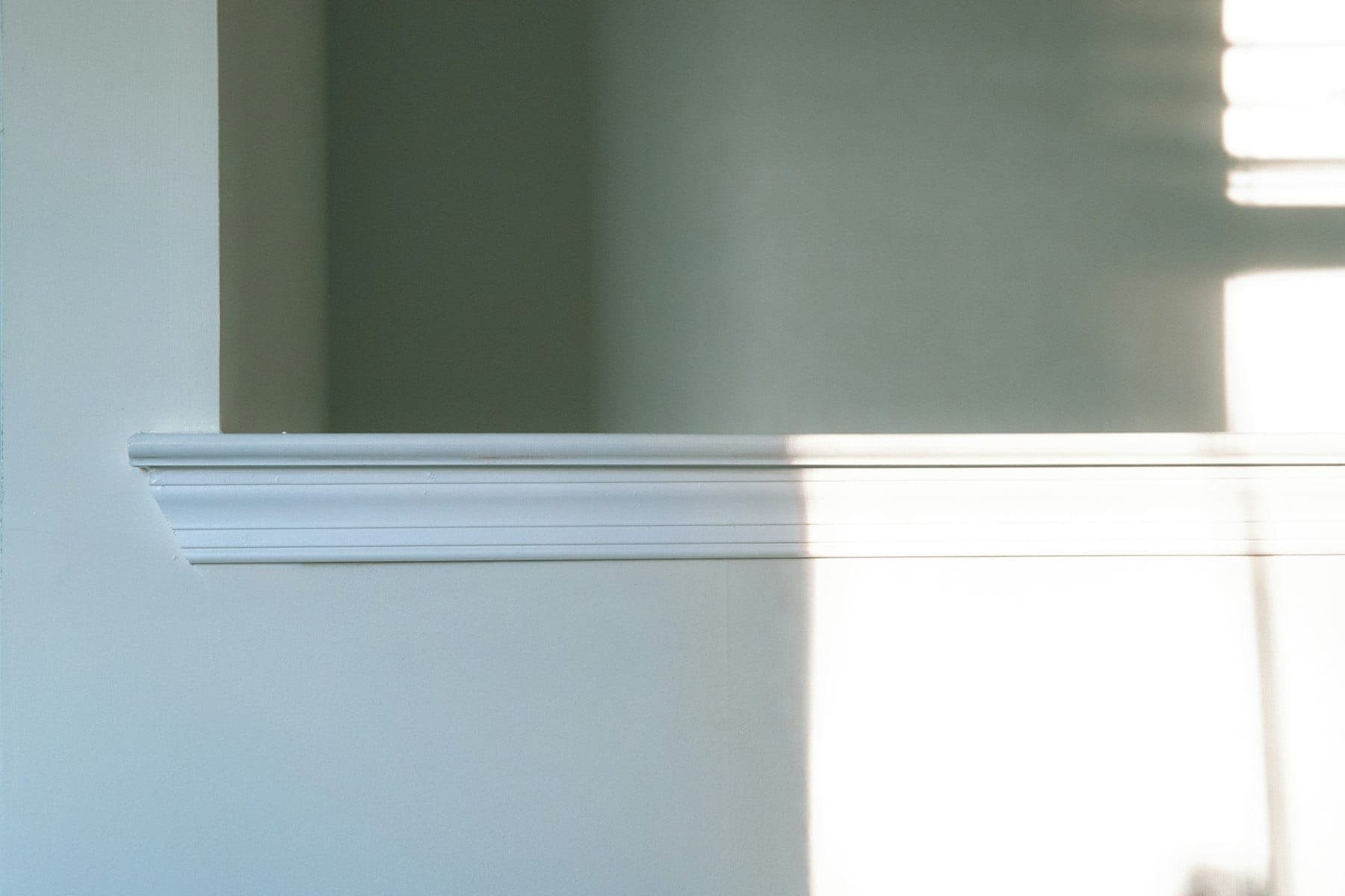 Crisp paint line where a sage-green upper wall meets a white chair rail and pale lower wall, in window light