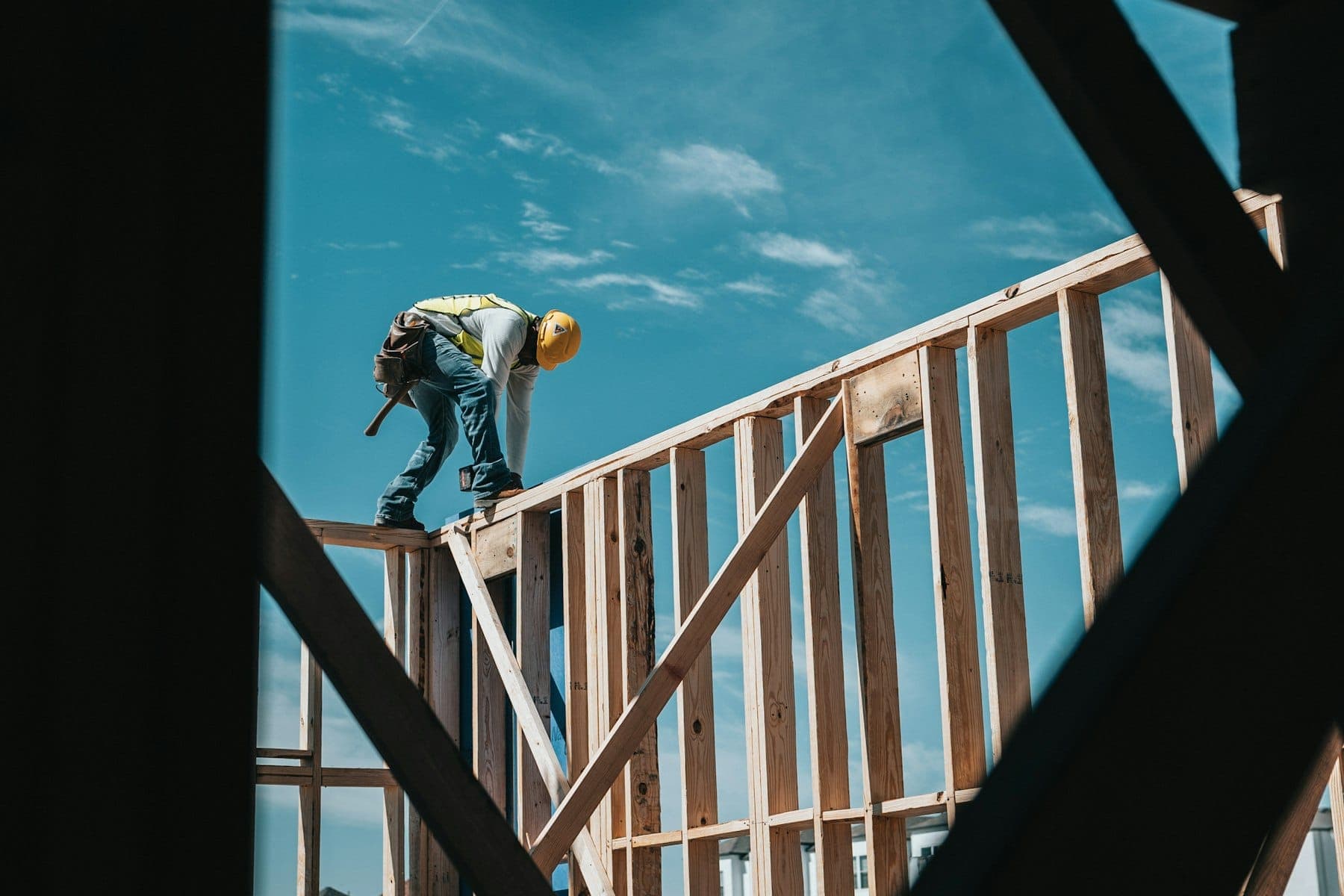 Framer in a hard hat and hi-vis vest working on top of a freshly framed wood wall, seen through the framing