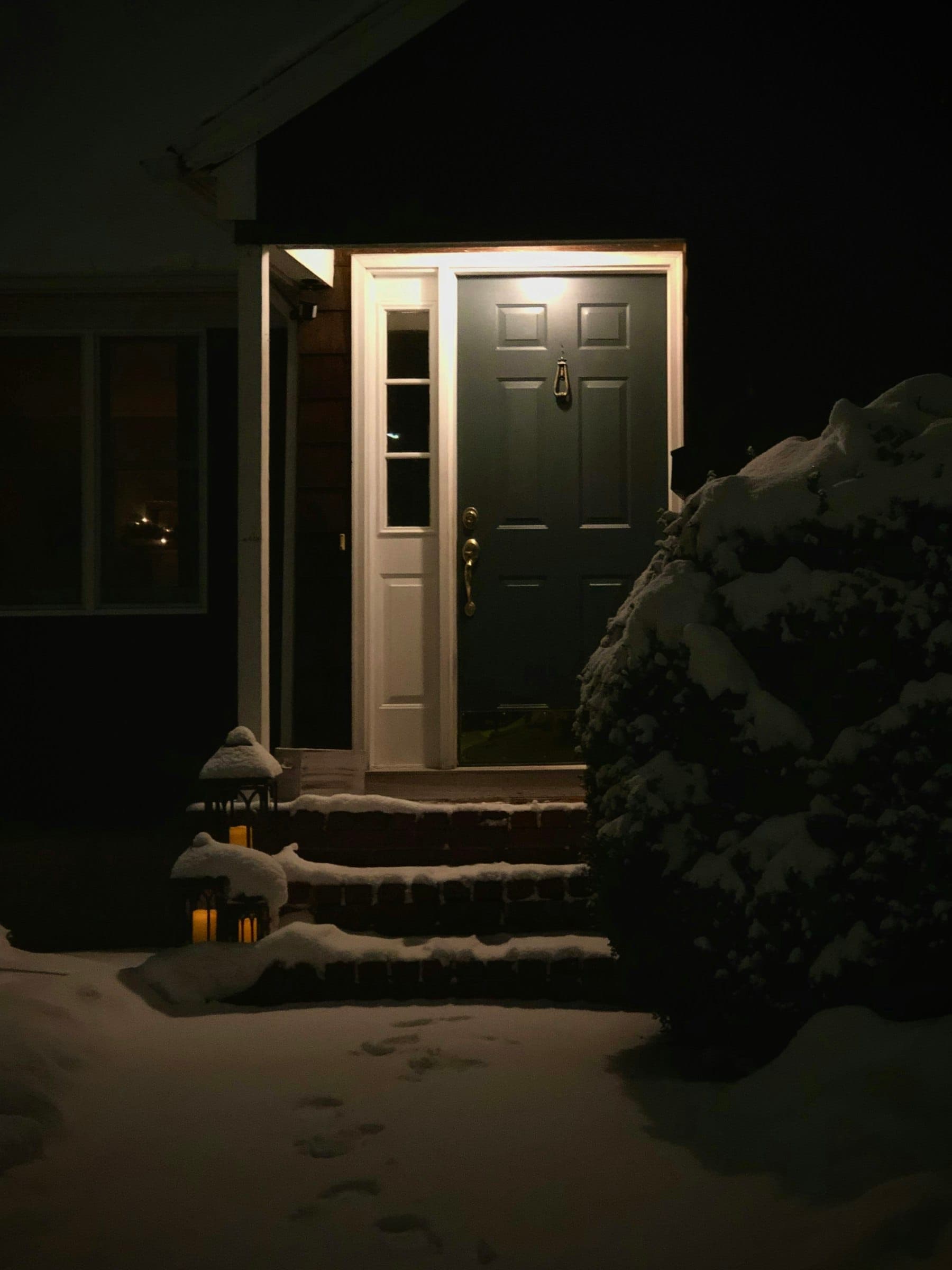 Dark-painted front door with a sidelight glowing under a porch light on a snowy night, footprints in fresh snow